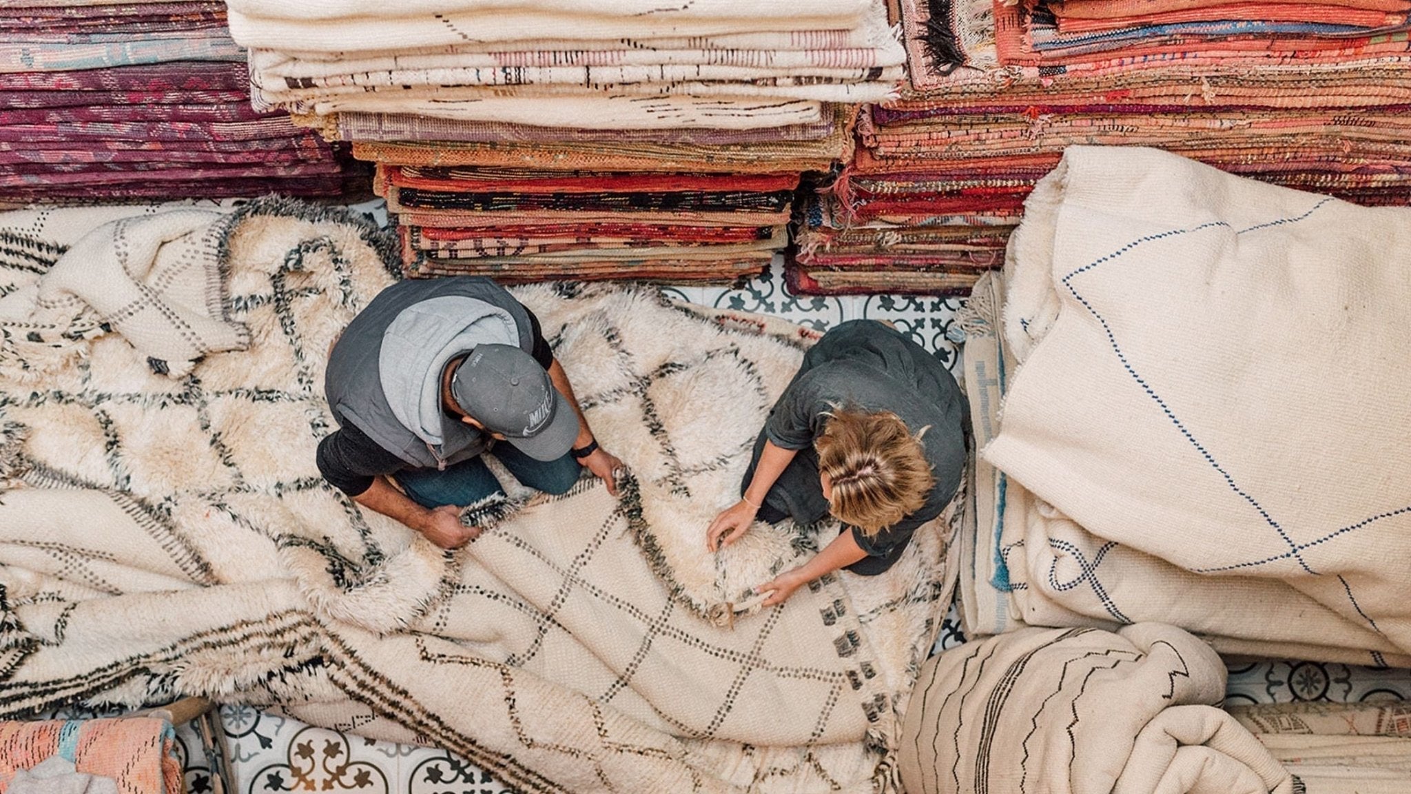 The Different Moroccan Rug Styles: Our Guide, 2 people sitting on a vintage Beni Ourain and stacks of rugs - Nouvelle Nomad