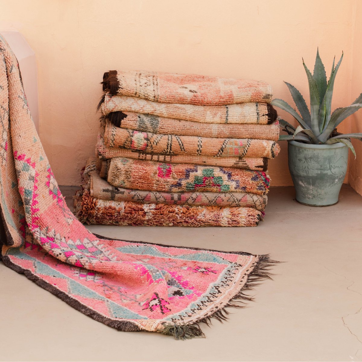 A stack of peach and pink coloured Moroccan rugs stacked and folded against a peach coloured wall | Nouvelle Nomad