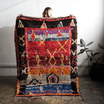 A person holds up a large, highly textured Moroccan-style wool rug with a vibrant multicolor geometric pattern, set against a white wall with a potted plant in the background.