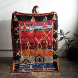 A person holds up a large, highly textured Moroccan-style wool rug with a vibrant multicolor geometric pattern, set against a white wall with a potted plant in the background.