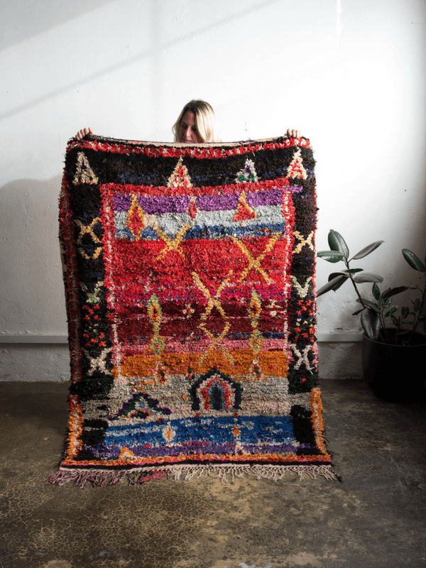 A person holds up a large, highly textured Moroccan-style wool rug with a vibrant multicolor geometric pattern, set against a white wall with a potted plant in the background.