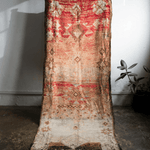 A vintage-style distressed area rug with a red, beige, and brown geometric pattern, draped vertically against a white wall in a room with a concrete floor and a small potted plant.
