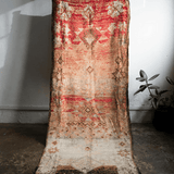 A vintage-style distressed area rug with a red, beige, and brown geometric pattern, draped vertically against a white wall in a room with a concrete floor and a small potted plant.