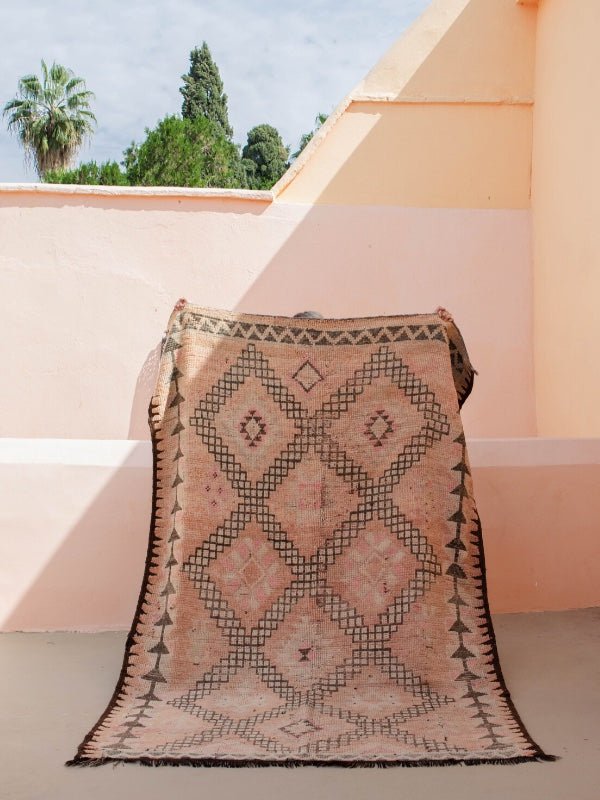 A soft faded pink vintage Moroccan rug being held up against a pink and peach wall - Nouvelle Nomad