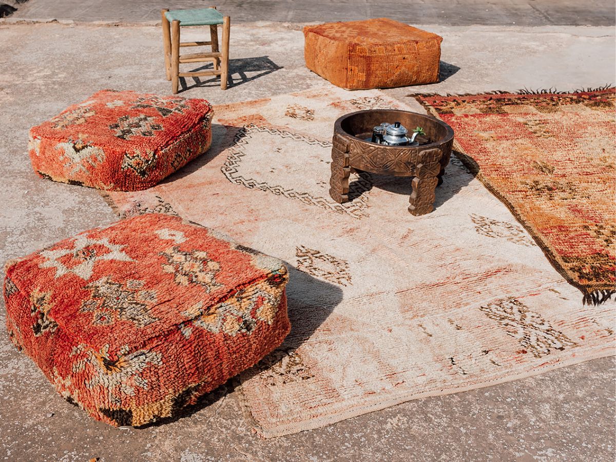 A faded pink Boujad rug on the ground with red Moroccan floor cushions and hand carved table