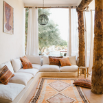 A bohemian-style living room featuring a built-in cream sectional sofa with patterned accent pillows, a central orange and brown geometric area rug, rustic exposed wood columns, and a large window looking out onto green trees.