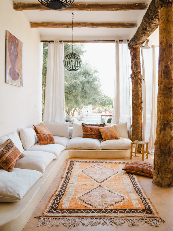 A bohemian-style living room featuring a built-in cream sectional sofa with patterned accent pillows, a central orange and brown geometric area rug, rustic exposed wood columns, and a large window looking out onto green trees.