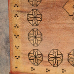 A close-up of a vintage-style rug corner showing a warm tan and orange-brown field with black geometric motifs and fringed edges.