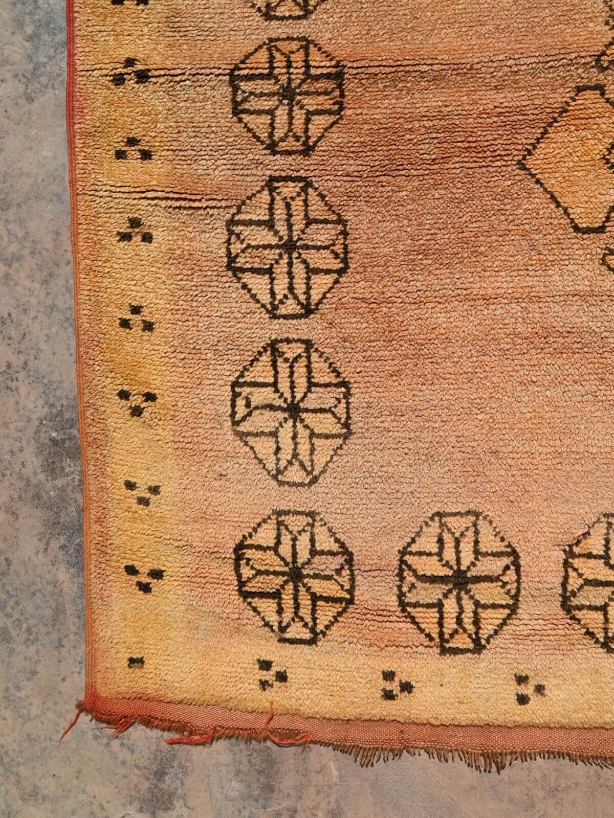 A close-up of a vintage-style rug corner showing a warm tan and orange-brown field with black geometric motifs and fringed edges.