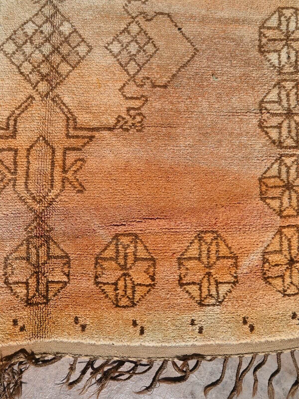 A close-up of a vintage-style woven rug with a warm tan and brown color palette, featuring geometric patterns and fringed edges.