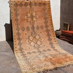 A large rectangular woven rug or textile panel with fringe, displayed hanging against a wall in a rustic indoor setting. The textile has a warm tan and beige base with dark brown geometric motifs, including repeated oval-and-cross patterns around the border and larger central medallion designs.