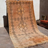 A large rectangular woven rug or textile panel with fringe, displayed hanging against a wall in a rustic indoor setting. The textile has a warm tan and beige base with dark brown geometric motifs, including repeated oval-and-cross patterns around the border and larger central medallion designs.