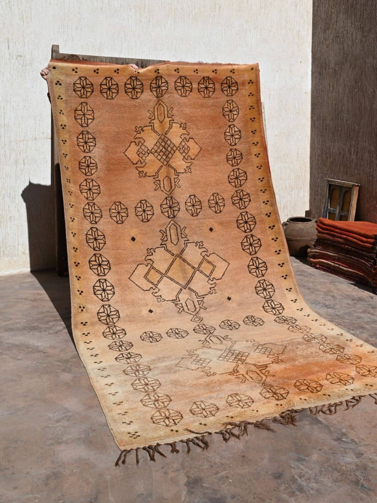 A large rectangular woven rug or textile panel with fringe, displayed hanging against a wall in a rustic indoor setting. The textile has a warm tan and beige base with dark brown geometric motifs, including repeated oval-and-cross patterns around the border and larger central medallion designs.
