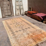 A rectangular vintage-style Moroccan rug with a faded tan and orange-brown field and dark brown geometric patterns, displayed on a stone floor in a rustic room with wooden doors and stacked textiles.