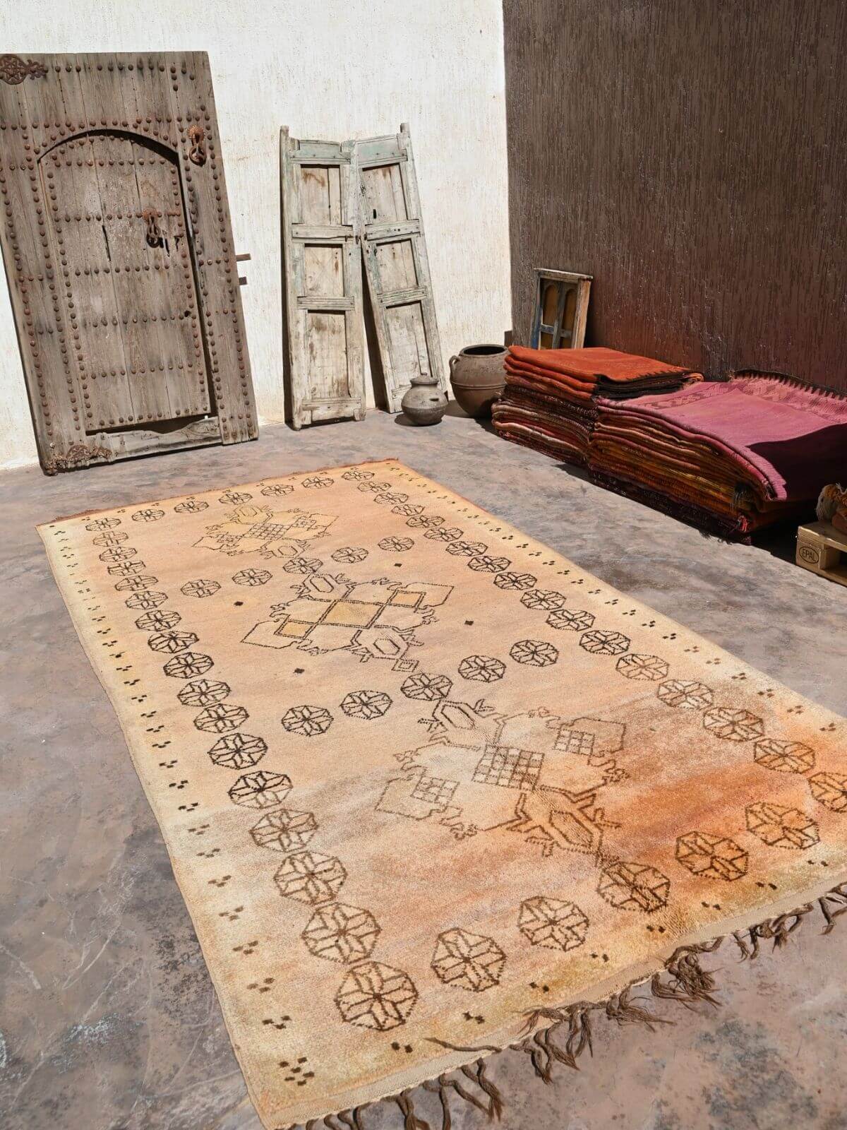 A rectangular vintage-style Moroccan rug with a faded tan and orange-brown field and dark brown geometric patterns, displayed on a stone floor in a rustic room with wooden doors and stacked textiles.