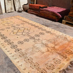 A large vintage Moroccan-style area rug with a faded ochre and beige wash, featuring geometric patterns and fringed edges, displayed on a rustic stone floor.