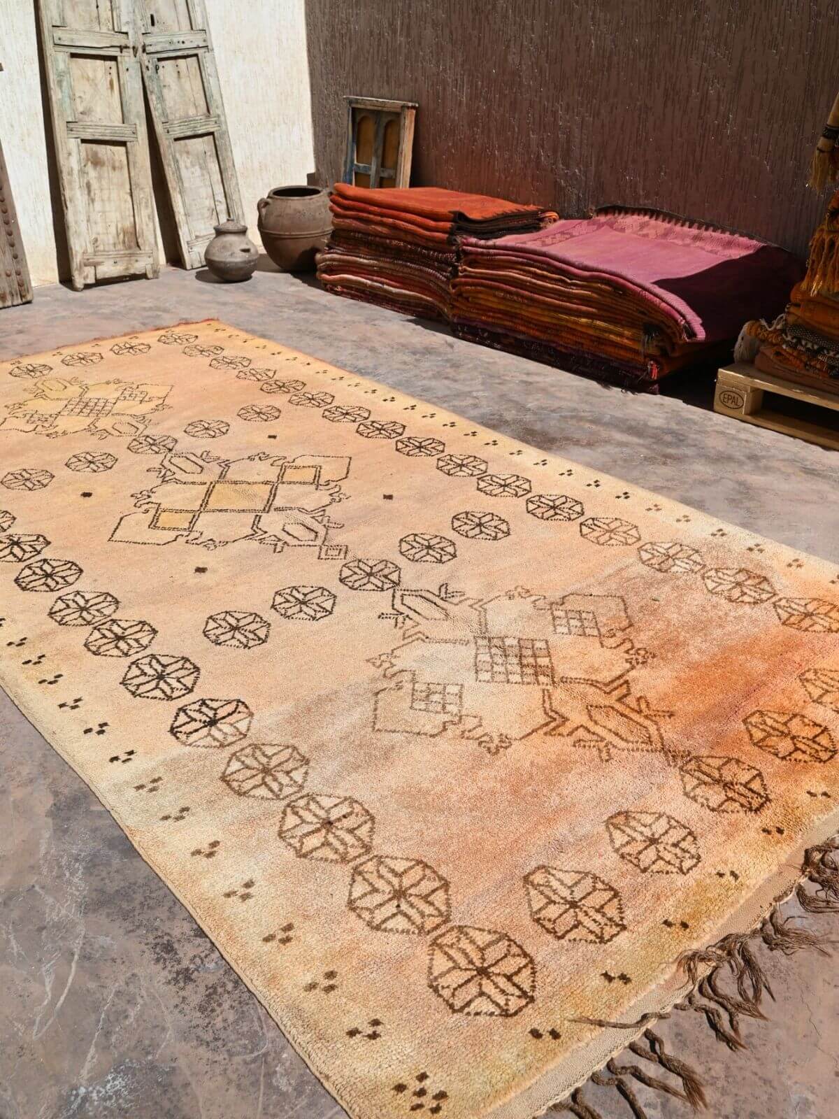 A large vintage Moroccan-style area rug with a faded ochre and beige wash, featuring geometric patterns and fringed edges, displayed on a rustic stone floor.